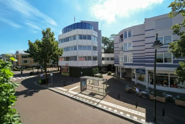 Straatbeeld van de 1e Hogeweg met moderne kantoorgebouwen, winkels, een bushalte en terras onder een blauwe lucht.